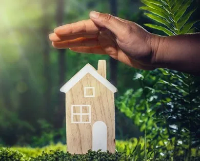 A hand sheltering a small wooden house model surrounded by lush green nature.