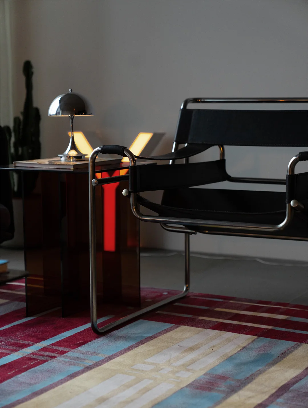 A Wassily-style tubular steel chair sits on a multicolour striped rug beside a chrome dome table lamp.