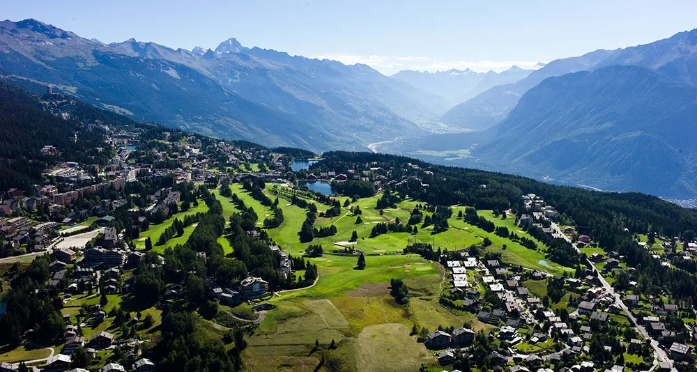 Aerial summer view of Crans-Montana showing the golf course, lakes, alpine village, and mountain valley.