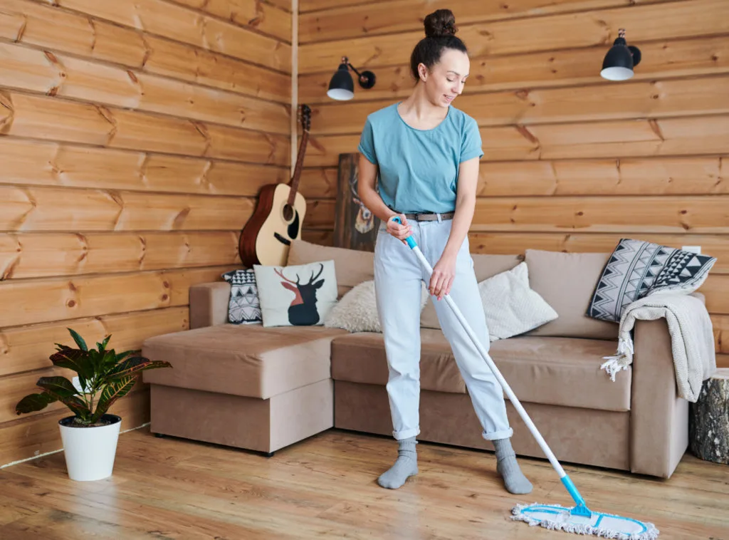 Woman mopping a rustic wooden-walled living room floor with a blue mop.