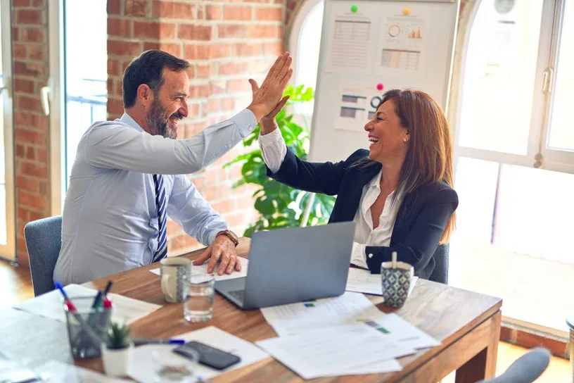 Two colleagues high-fiving at desk in bright office—captures the joy and partnership spirit central to Putzwind's founding story.