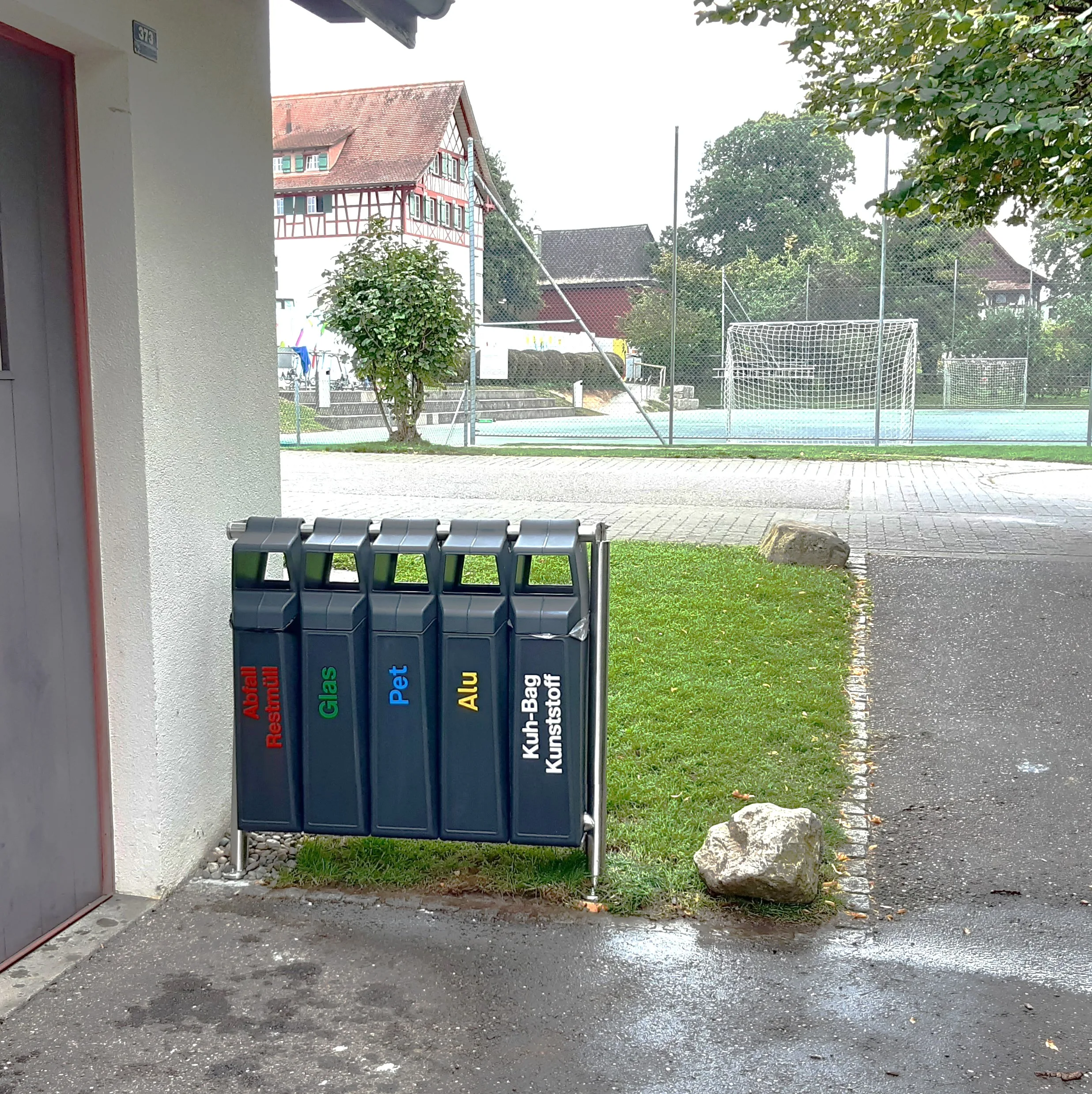 A five-compartment outdoor recycling station with labelled bins for waste sorting mounted near a sports field.