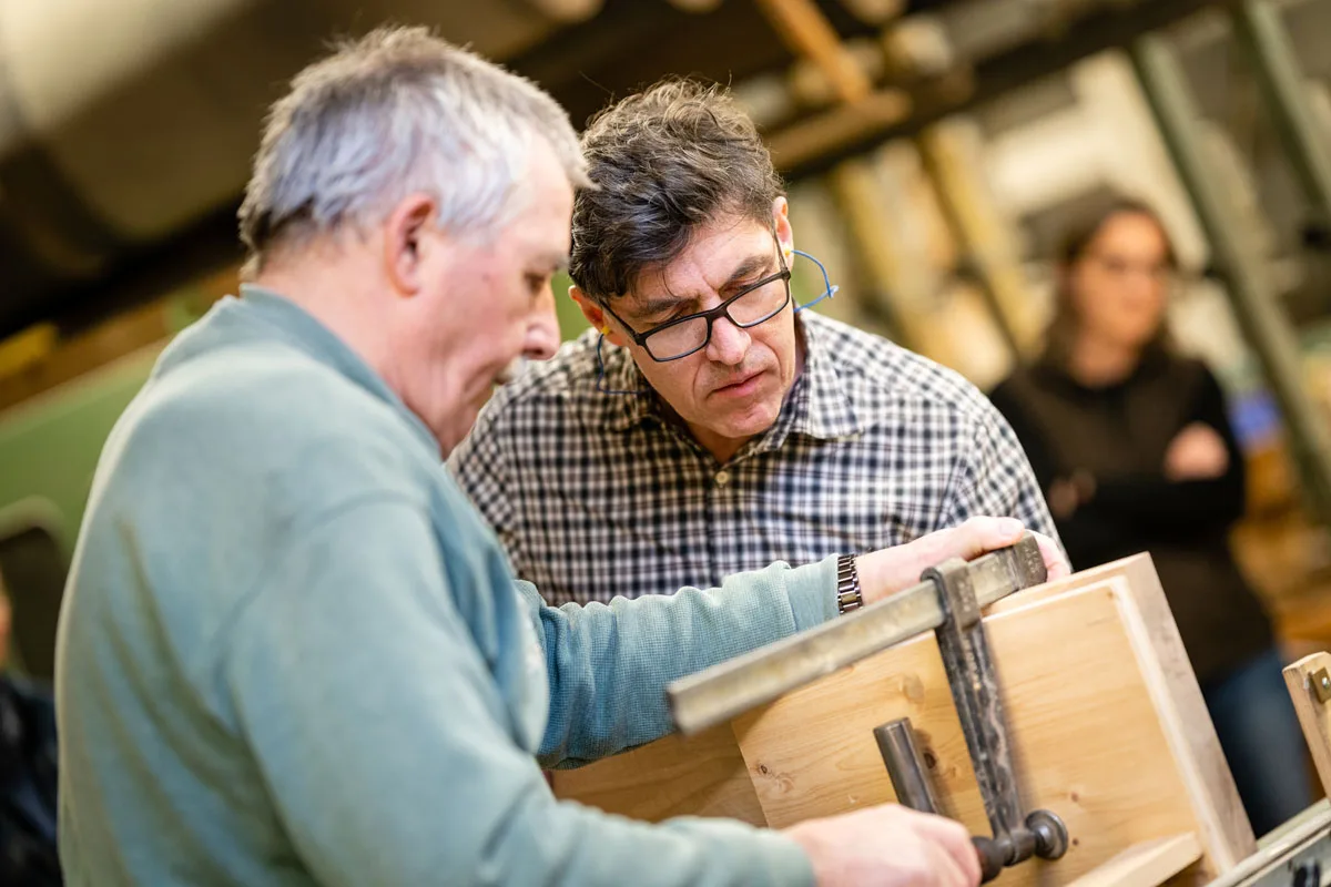 A skilled craftsman carefully inserts magnets into a drilled wood block beside a drill press.