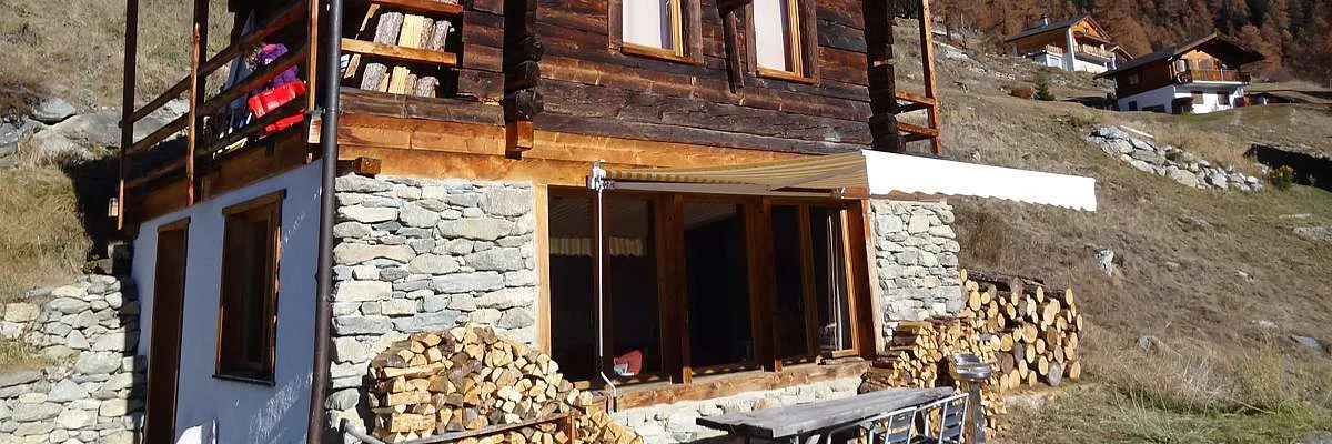 Dark weathered chalet with stone wall, firewood stacks, and awning in late-autumn mountain setting.