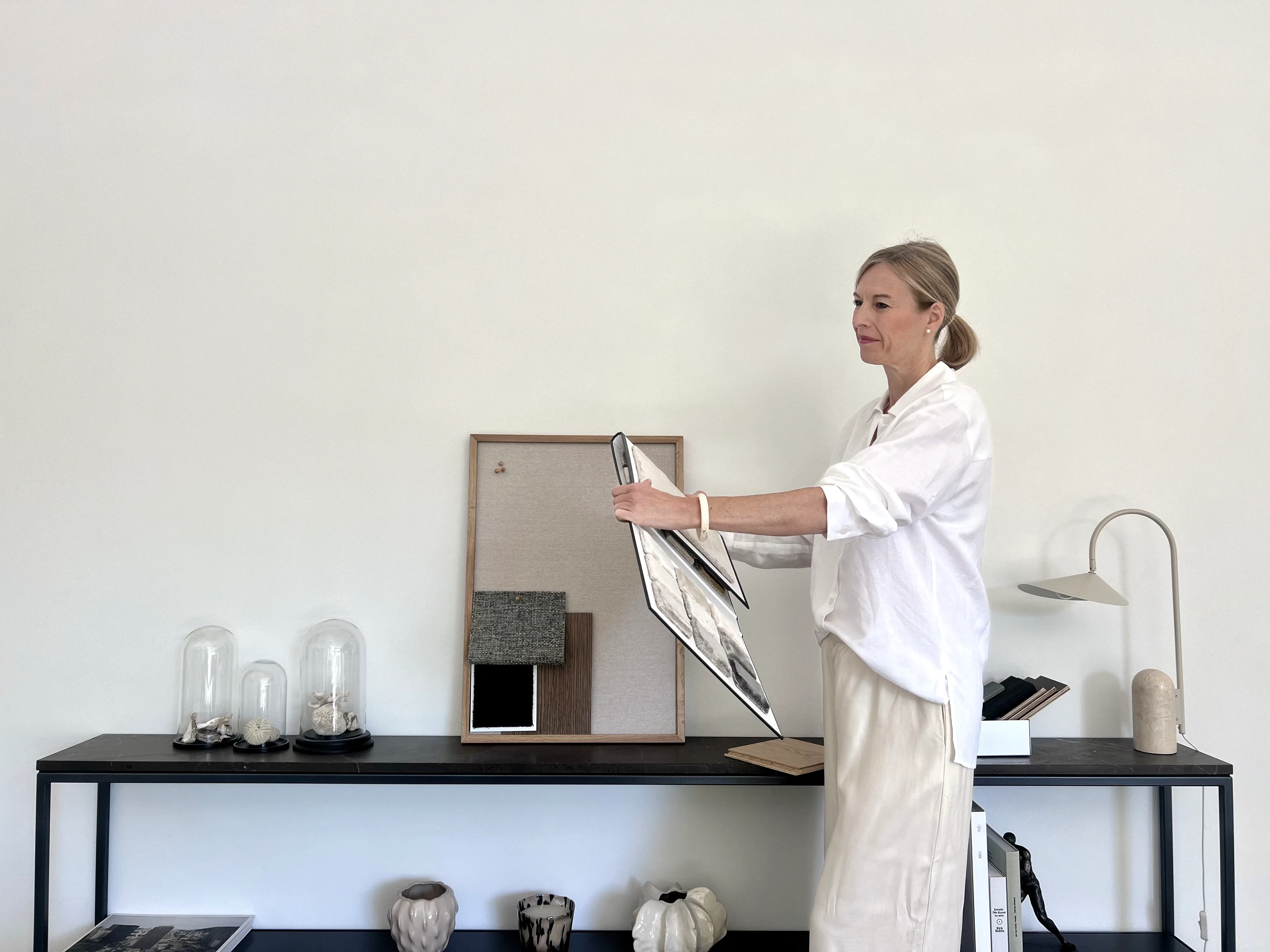 A woman in white linen arranges material samples on a sleek console in a bright studio.