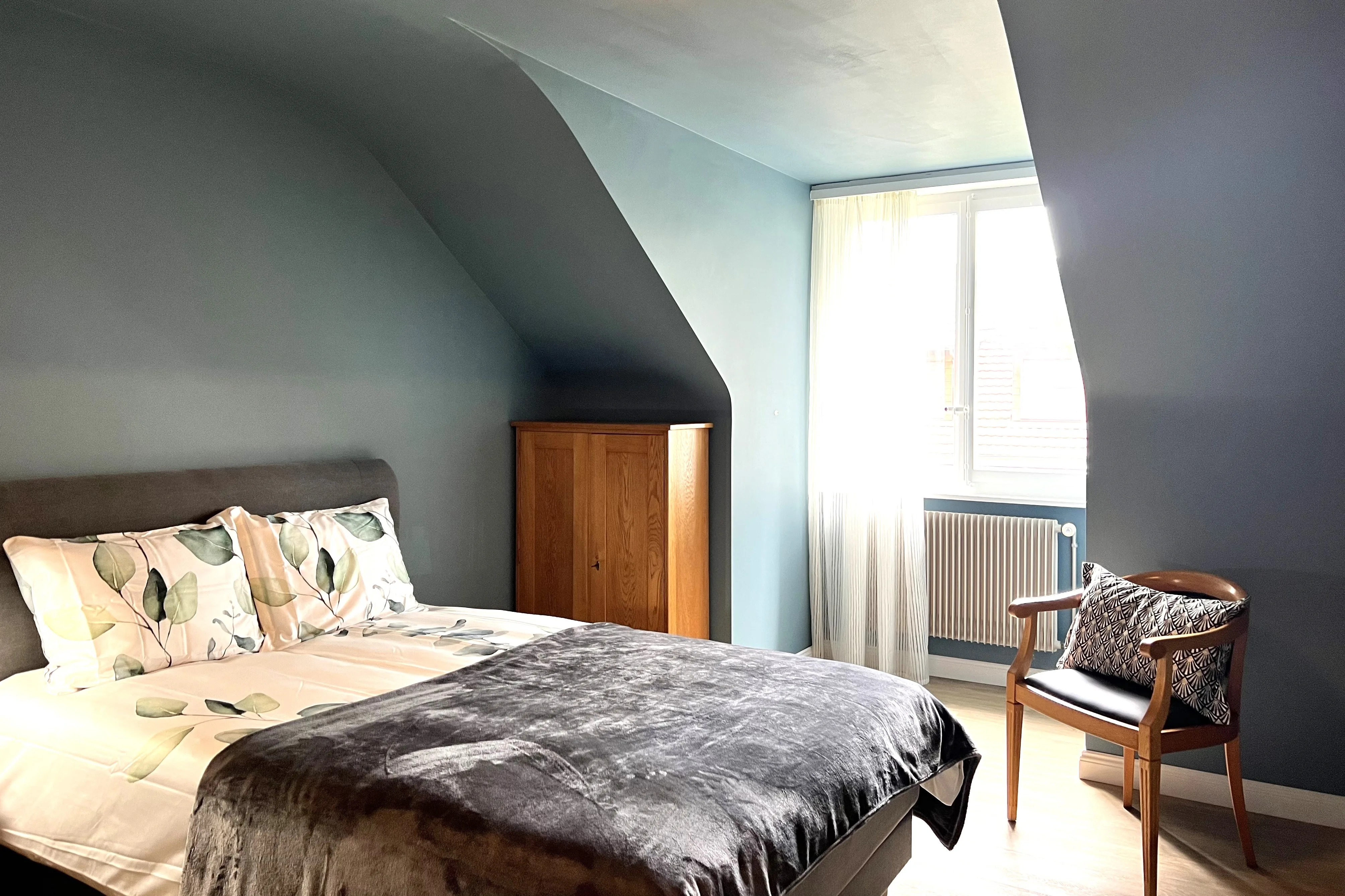 Cozy attic bedroom with blue-grey walls and layered textiles, showing Bless Interieur's signature use of color psychology to create intimate, welcoming spaces.